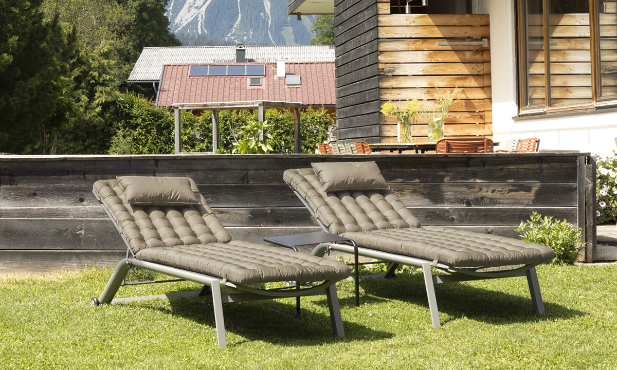 Zwei gepolsterte Gartenliegen mit kleinem Beistelltisch auf einer Wiese vor einem Holzhaus.